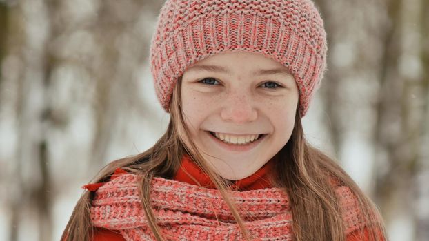 Portrait of a young schoolgirl with freckles in the woods in winter. He warms his hands in mittens and applies them to his face and lips. Shows movement hand in hand