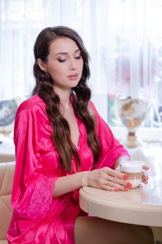 morning routine. charming young brunette woman sitting in her kitchen in pink dressing gown or robe with a cup of coffee. tender woman portrait.