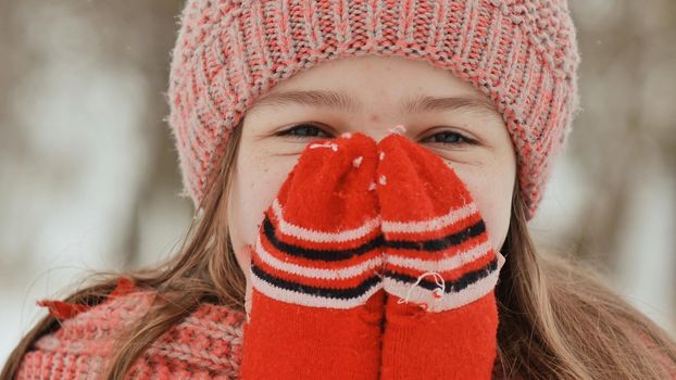 Portrait of a young schoolgirl with freckles in the woods in winter. He warms his hands in mittens and applies them to his face and lips. Shows movement hand in hand