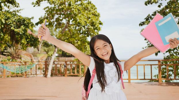 A charming philippine schoolgirl with a backpack and books in a park off the coast. A girl joyfully poses, raising her hands up with textbooks in her hands. Warm sunny day