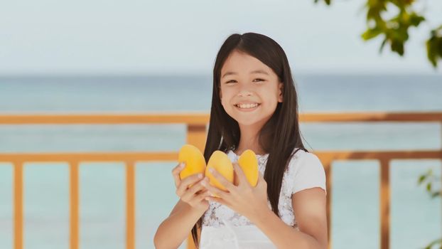 A charming philippine schoolgirl girl in a white dress and long hair positively poses with a mango in her hands. The sun. The blue ocean. Childhood. Recreation