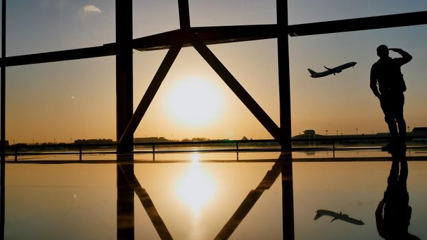 Silhouette of a tourist guy watching the take-off of the plane standing at the airport window at sunset in the evening. Travel concept, people in the airport
