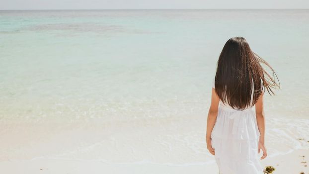 A charming philippine schoolgirl in a white dress is walking along a white sandy beach. Enjoying the tropical scenery. Childhood