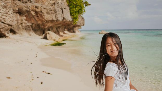 A charming and happy philippine teenage girl in a white summer dress is running along a tropical beach near the rocks. She is happily spinning. Childhood. Recreation