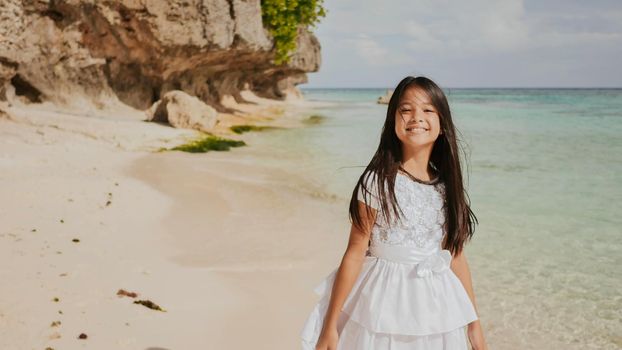 A charming and happy philippine teenage girl in a white summer dress is running along a tropical beach near the rocks. She is happily spinning. Childhood. Recreation