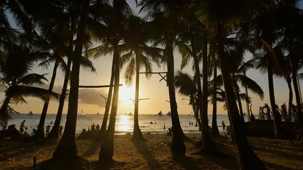 Beach with silhouettes of tourists among palm trees on the island of Boracay. Palm trees in the rays of sunset. Sailboats on the water. Philippine Tropics