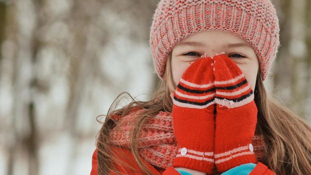 Portrait of a young schoolgirl with freckles in the woods in winter. He warms his hands in mittens and applies them to his face and lips. Shows movement hand in hand