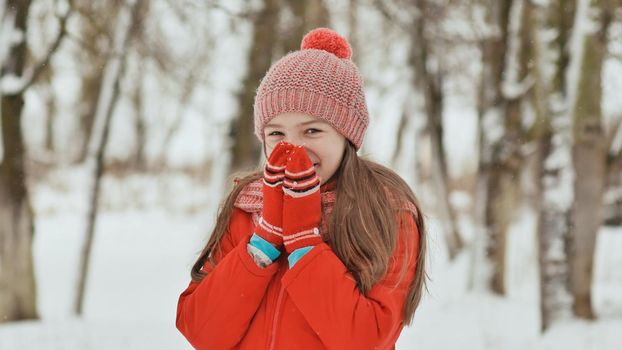 Portrait of a young schoolgirl with freckles in the woods in winter. He warms his hands in mittens and applies them to his face and lips. Shows movement hand in hand
