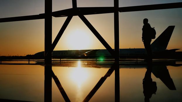 The silhouette of the tourist guy watching the takeoff of the plane and passing a huge plane standing at the airport window at sunset in the evening. The travel concept, people in the airport