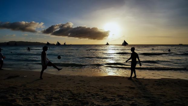 Beach on the island of Boracay in the rays of the evening sunset. Silhouettes of people playing a ball on the beach and sailboats on the water