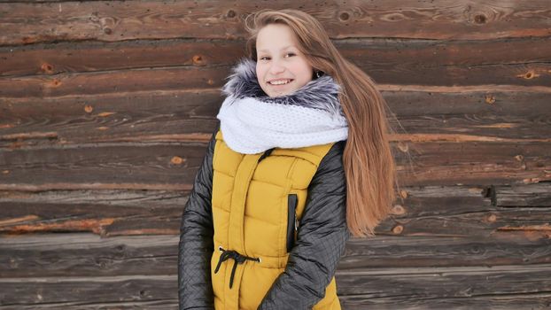 Young beautiful girl in winter clothes posing positively at the camera on the background of a wooden house in the village