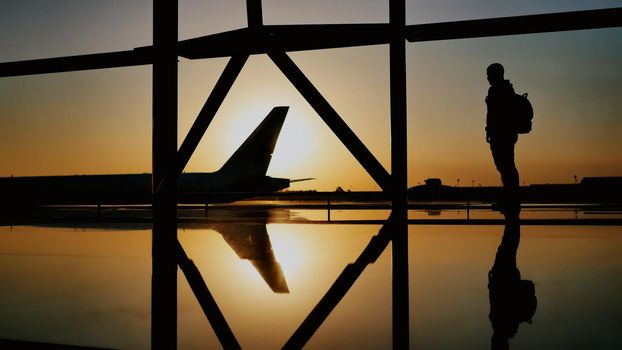 The silhouette of the tourist guy watching the takeoff of the plane and passing a huge plane standing at the airport window at sunset in the evening. The travel concept, people in the airport