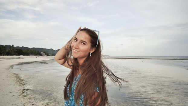 A beautiful girl with long hair in a long blue dress strolls along the beach. Girl posing on camera on exotic coast. Shooting in motion