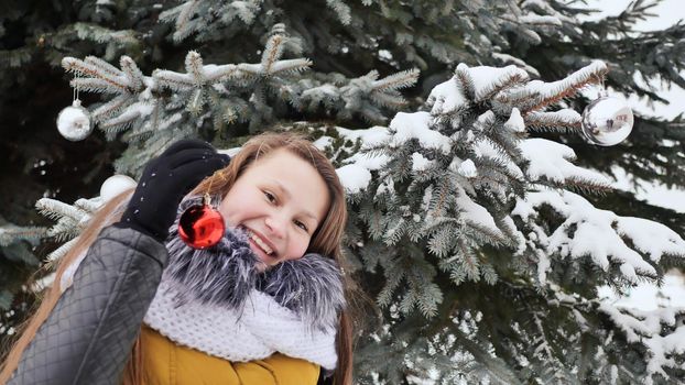 Attractive young girl with long hair in a winter suit posing against a snowy tree. A girl is stringing Christmas balls on a branch. Winter in the forest