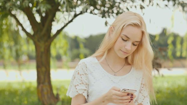 A young blonde girl is typing on white phone in a city park.