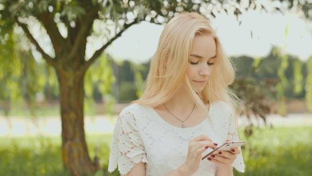 A young blonde girl is typing on white phone in a city park.