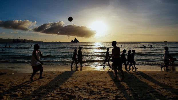 Beach on the island of Boracay in the rays of the evening sunset. Silhouettes of people playing a ball on the beach and sailboats on the water