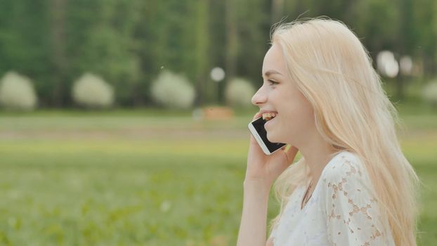 A young blonde girl is talking on the phone in a city park sitting on the grass