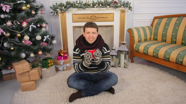 A young guy sits on the floor of his house with a mug of hot tea. Christmas theme