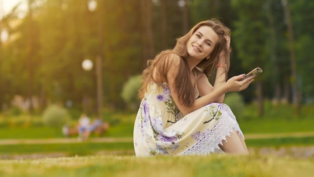 Young beautiful woman in summer dress with long hair sitting on grass in green park and talking on the phone, smiling. Summer. Recreation. Youth