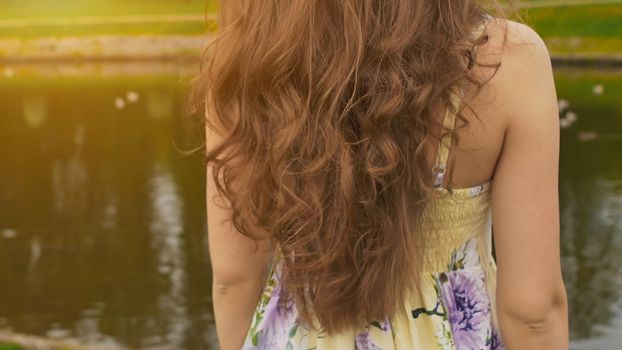 A girl with long, beautiful and curly hair against the backdrop of a pond in the park. Hair close-up. The girl sensually touches her hair. Youth. Beauty. Summer