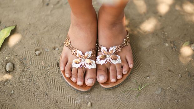 Tanned legs of a little girl in slates with a decorative butterfly. Girl barefoot in summer shoes on sand. Philippines