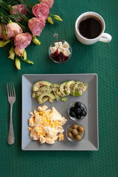 Nutritious breakfast per person: scrambled eggs, olives, olives, avocados, cheese and raspberry jam. Next to it is a cup of black coffee and a bouquet of flowers.