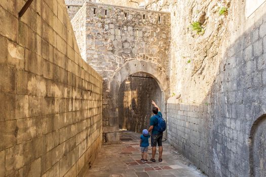 Father and son walking among medieval fortress walls in Kotor city, Montenegro. Tourists exploring narrow paved stone street in the old town on a summer sunny day.