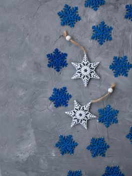 Two white ornaments on a Christmas tree in the form of snowflakes and many blue snowflakes lie on a gray concrete background. There is a place for a welcome text.
