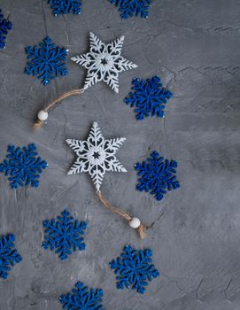 Two white ornaments on a Christmas tree in the form of snowflakes and many blue snowflakes lie on a gray concrete background. There is a place for a welcome text.