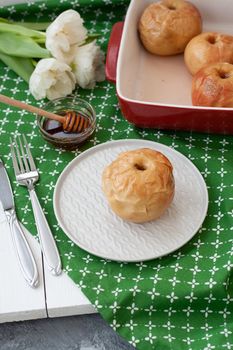 Baked apples in red, honey, a bunch of white tulips lie on a green tablecloth.