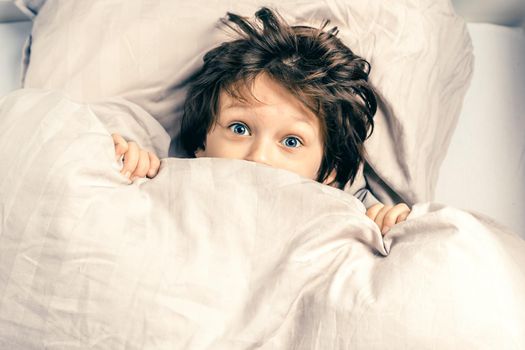 Surprised boy with funny face expression hiding and peeking from a duvet. Kid covering half of his face with blanket. Scared child, afraid of night monsters