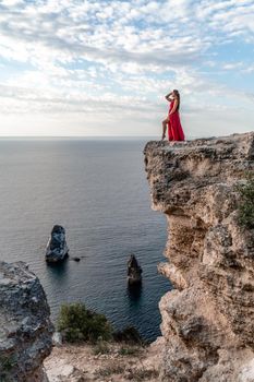 A girl with loose hair in a red dress stands on a rock rock above the sea. In the background, the sea and the rocks. The concept of travel.