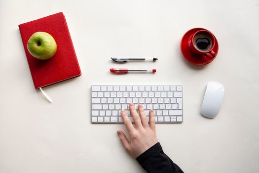 Man hands on wireless keyboard with red notebook, green apple and red cup of coffee on white table