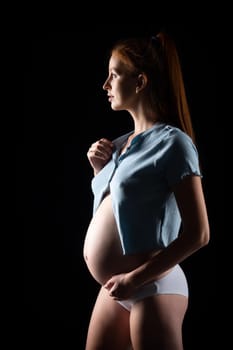 A pregnant woman with a big belly and red hair. Posing on a black background in the studio.