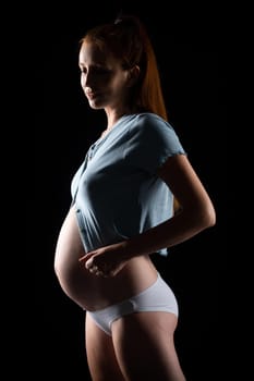 A pregnant woman with a big belly and red hair. Posing on a black background in the studio.