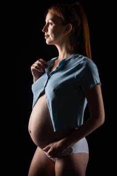 A pregnant woman with a big belly and red hair. Posing on a black background in the studio.