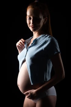 A pregnant woman with a big belly and red hair. Posing on a black background in the studio.