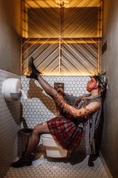 a punk girl sitting on the toilet straightens her torn black stockings. The girl has a stocking mask on her head.