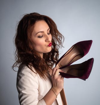 girl in a beige jacket and shoes in her hands, posing for the camera. Shot in a photo studio on a gray background.