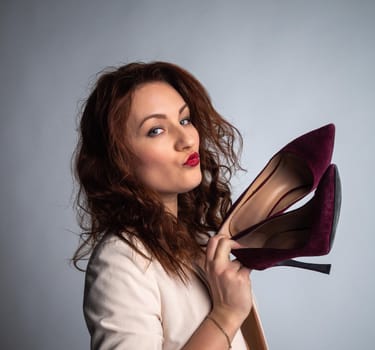 girl in a beige jacket and shoes in her hands, posing for the camera. Shot in a photo studio on a gray background.