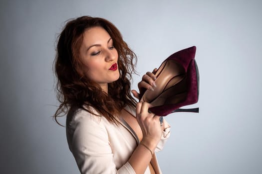 girl in a beige jacket and shoes in her hands, posing for the camera. Shot in a photo studio on a gray background.