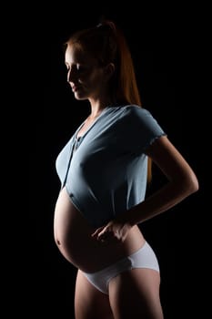 A pregnant woman with a big belly and red hair. Posing on a black background in the studio.