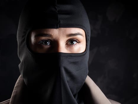 Portrait of a girl in a black balaclava and beige coat. Shot in the studio on a dark background.