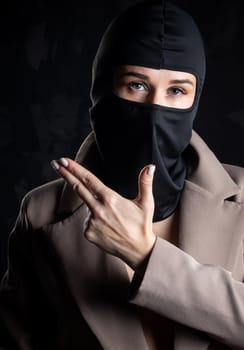 Portrait of a girl in a black balaclava and beige coat. Shot in the studio on a dark background.