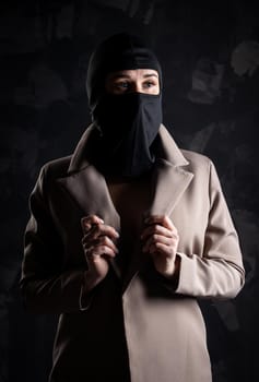 Portrait of a girl in a black balaclava and beige coat. Shot in the studio on a dark background.