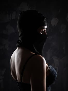 Portrait of a girl in a black balaclava and a black bra. Shot in the studio on a dark background.