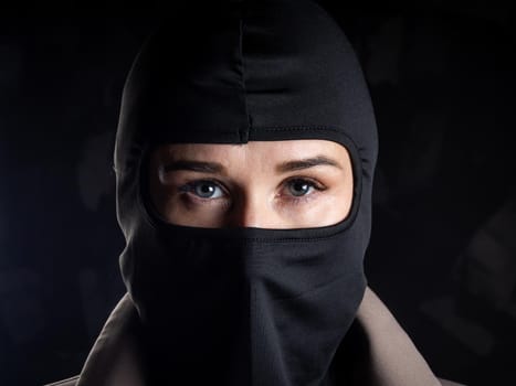 Portrait of a girl in a black balaclava and beige coat. Shot in the studio on a dark background.