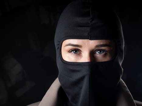 Portrait of a girl in a black balaclava and beige coat. Shot in the studio on a dark background.