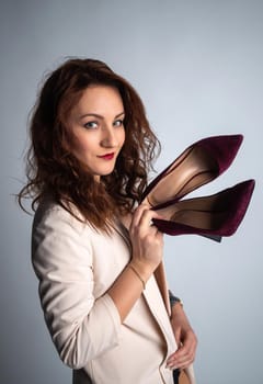 girl in a beige jacket and shoes in her hands, posing for the camera. Shot in a photo studio on a gray background.